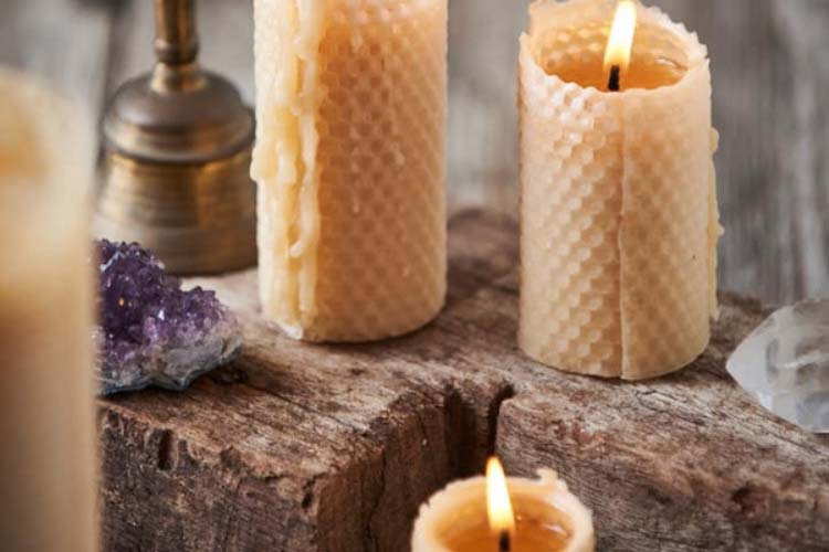 A zoomed in photo of beeswax candles resting on a thick wooden block. The candles has a honeycomb pattern stamped on the outside and a coloured in a golden/honey shade. Also resting in the surroundings are two crystals and a brass bell.
