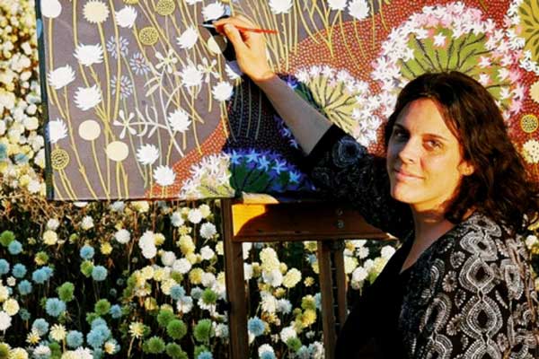 A photo of artist Helen Ansell, looking towards the viewer, with golden sunlight lighting up her face. Their right hand is holding a thin red paintbrush suspended over an upright canvas covered in painted flowers and a dot motif.In the background is a field of red white and yellow flowers, spherical in form.