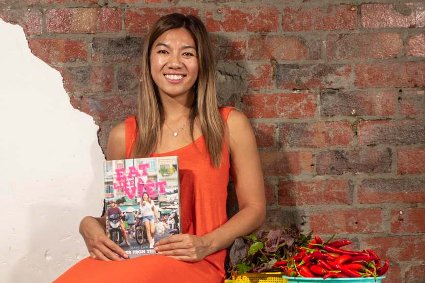 A portait of Jenny Lam in an orage sleeveless dress seated in front of an exposed brick wall. Jenny is holding up a copy of her book Eat like a Việt. Next to her are a tray of herbs and a tray of red chillies.