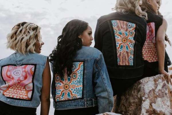 A photo of four models sitting on rocks with their backs turned towards the camera. They are wearing blue and black denim jackets with back panels made of colourful dot pattern artwork.