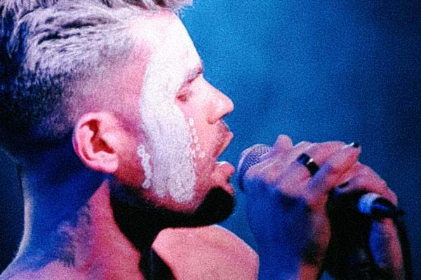 A close up photo of artist Boox Kid performing on stage. His hands are wrapped around the microphone and his face turned towards the right. He has short cropped hair and facepaint running from his cheek to his forehead.