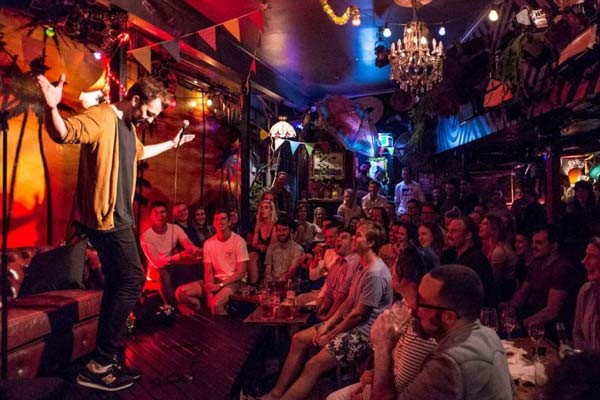 A photo of a comedy show at a bar. On the right and flanking the stage is a seated crowd reacting to the comedian. On the left is the comedian standing stage, behind a microphone, with their arms streched out and head lowered suggesting acceptance of the audience reaction. The bar decor is hippie-bohemian with a wallpaper of a palm beach sunset scene, a crystal chandelier, parasols, lampshades, and coloured flags hanging off the walls and ceiling.