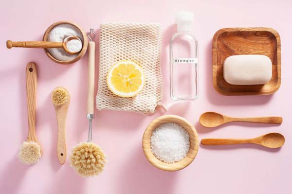 A top down photo of a light pink coloured surface with various wooden tools, utensils, and cleaning materials. Clockwise from the top is a small wooden bowl with a rounded spoon dipped in a white powder, a white folded netted cloth with a half of a lemon, a bottle labelled vinegar, a square wooden tray with a bar of soap, two wooden tea spoons, a small wooden bowl containing a flaky white substance, and finally three wooden scrubbing brushes in varying sizes.