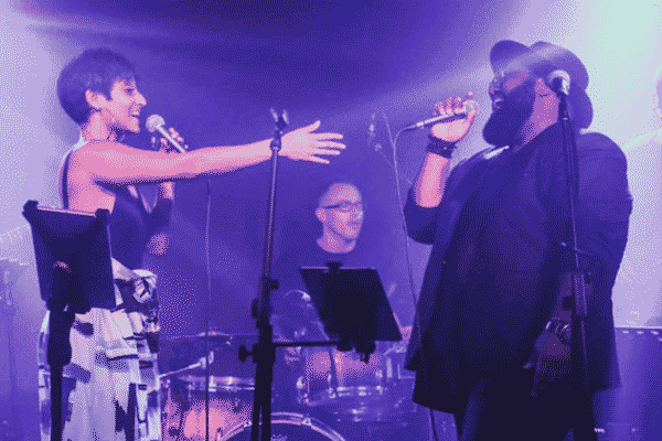 A photo of Elise Lynelle and Solomon Pitt performing on stage. Elise is seen reaching a hand out towards Solomon. A drummer can be seen between them in the background.