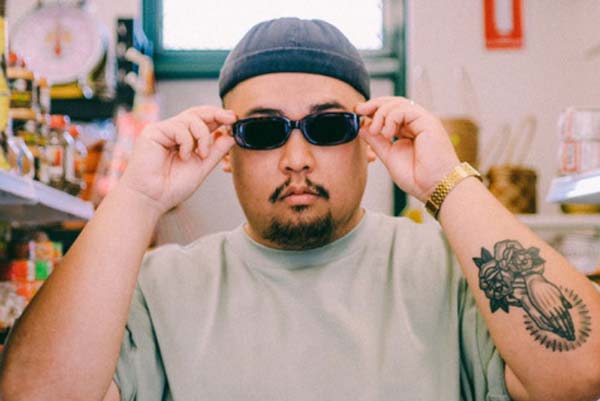 A portait of the artist Rein, standing in an shop aisle, looking towards the camera. They are wearing a brim-less cap, a light sage coloured tee shirt, a gold watch, and rectangular sunglasses. Their hands are holding the temples of the sunglasses. On their left arm is a tattoo of hands held in prayer with two roses below and a halo around.