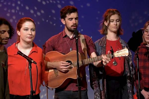 A photo of five members of the Koondarm choir performing on stage. They are dressed in red and black, standing against a purple backgdrop. The performer in the center of the frame is playing an acoustic guitar and singing in to a microphone.