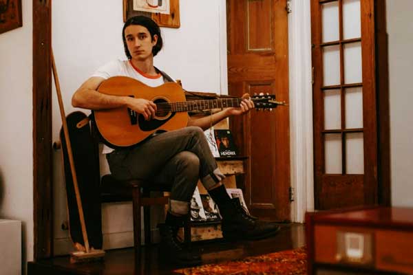 A full body portrait of Jack Davies seated on a chair with an acoustic guitar in hand. Seated in what looks to be a hallway, flanked by a broom, skateboard, and a bookshelf.