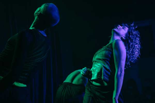 A photo of three performers on stage, bent backwards and heads tilted upwards almost in a trance. The left side of their bodies are lit by a green stage light while the right is lit purple.