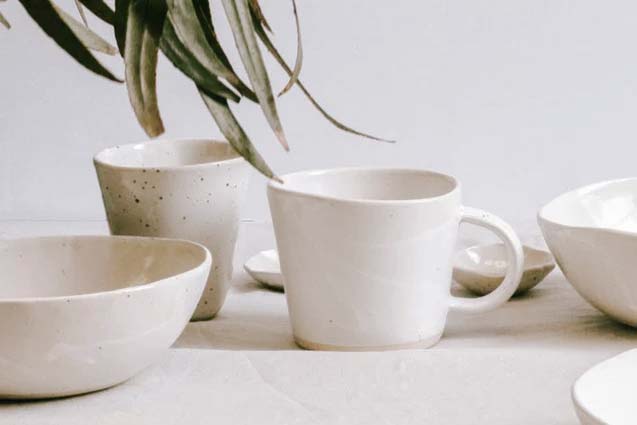 A photo of a white table with various ceramic vessels (3 bowls, a cup, a mug, and two mini dishes) all glazed white. Creeping in from the top right is a bunch of Eucalyptus leavs.
