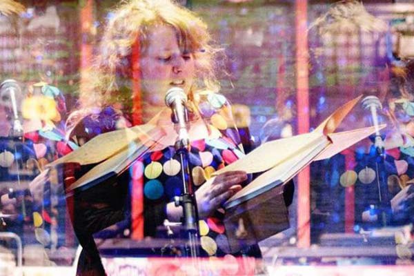 A photo of Saoise Nash, a person with long red hair wearing a dress adorned with coloured circles of blue, white, red, and yellow, holding a book and speaking in to a microphone. The image has be manipulated to show faint repetitions of the subject on the left and right.